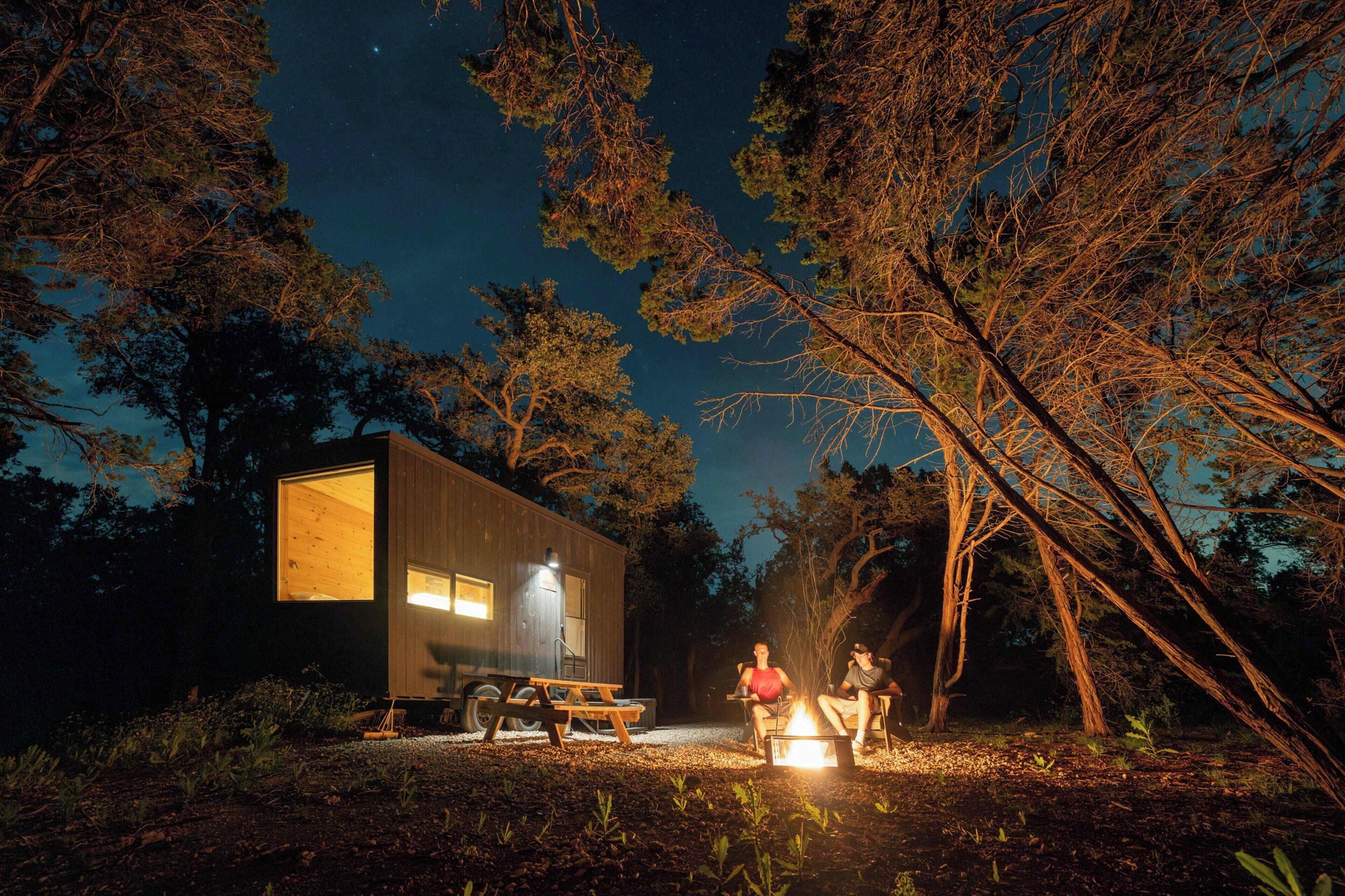 A modern cabin with a large window, a picnic table, and a campfire.