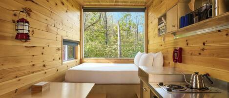 A cabin interior with a wooden interior, a bed, a dining area, a kitchen with a stove, and a window with a view of trees.