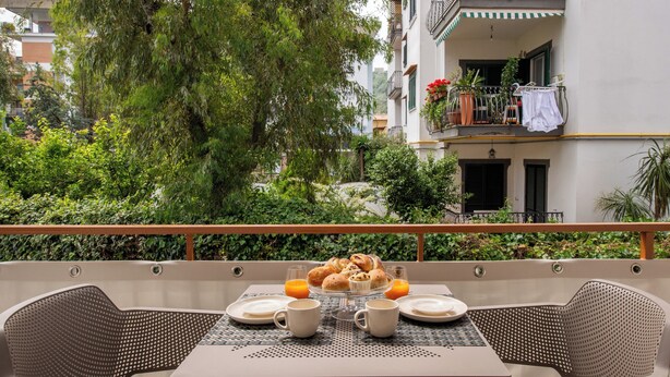 An outdoor dining area with wicker chairs, a table set with plates, cups, and a bowl of pastries, surrounded by greenery and a building with balconies. - Comfortable apartment in Sorrento center (Sorrento)