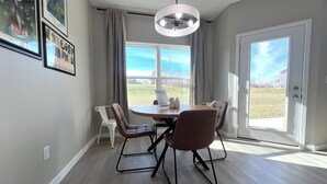 Comedor con una mesa redonda, cuatro sillas, un ventilador de techo y vista a un campo cubierto de hierba a través de un ventanal grande.