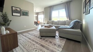 Una sala de estar con sofá, reposapiés y un tapiz estampado. En el fondo, se aprecia un comedor con mesa y sillas.