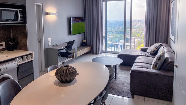 A modern living room with a dining area, a flat-screen TV, and a large window offering a city view. - Sandtoncity-apartment-sleeps2-pool-parking (Johannesburg)