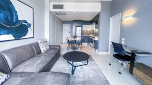 A modern living room with a grey sofa, a black coffee table, and a dining area with a table and chairs. A kitchen is visible in the background with a washing machine. - Sandtoncity-apartment-sleeps2-pool-parking (Johannesburg)