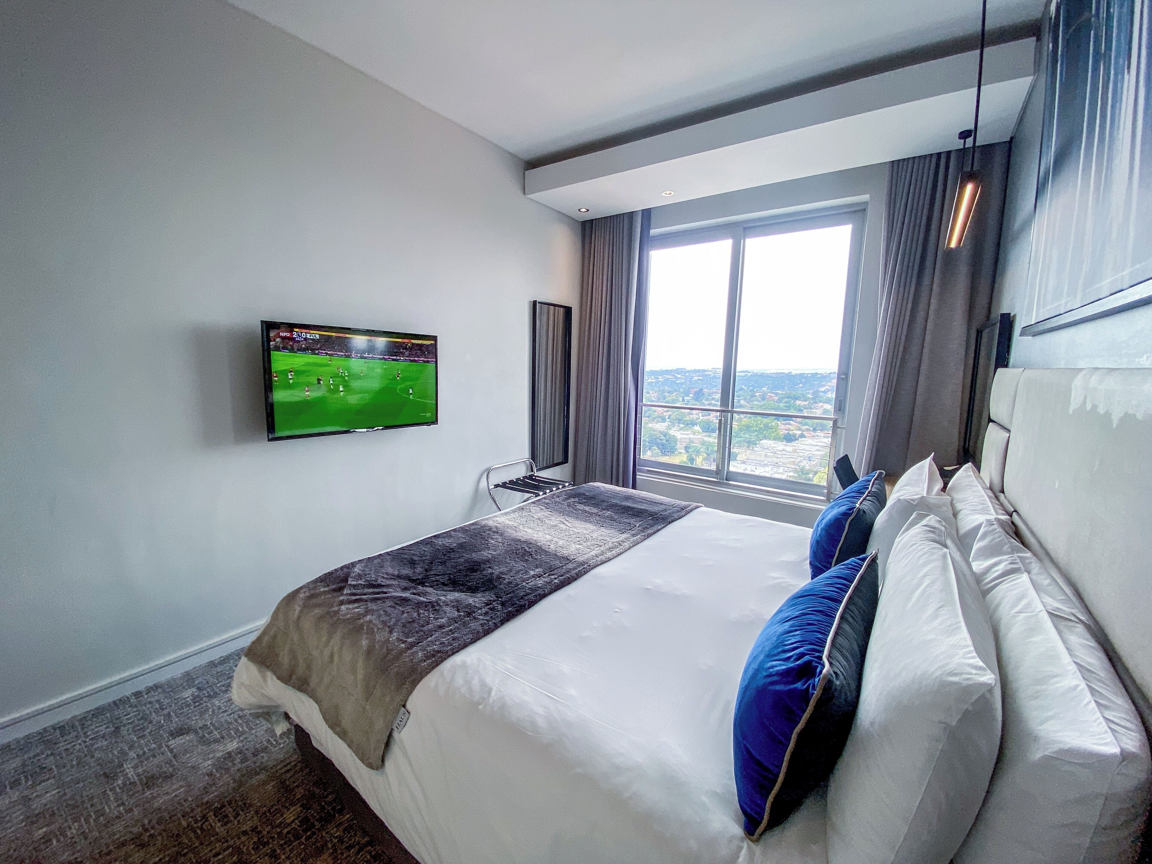 Una habitación de hotel con una cama grande, un televisor de pantalla plana montado en la pared, una ventana con vistas a la ciudad y un diseño moderno del techo.