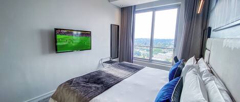 Una habitación de hotel con una cama grande, un televisor de pantalla plana montado en la pared, una ventana con vistas a la ciudad y un diseño moderno del techo.
