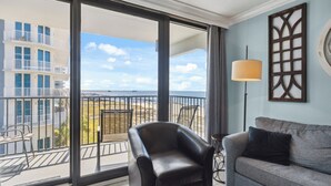 A balcony with a view of a building and beach, a black leather chair, a grey sofa, and a floor lamp.