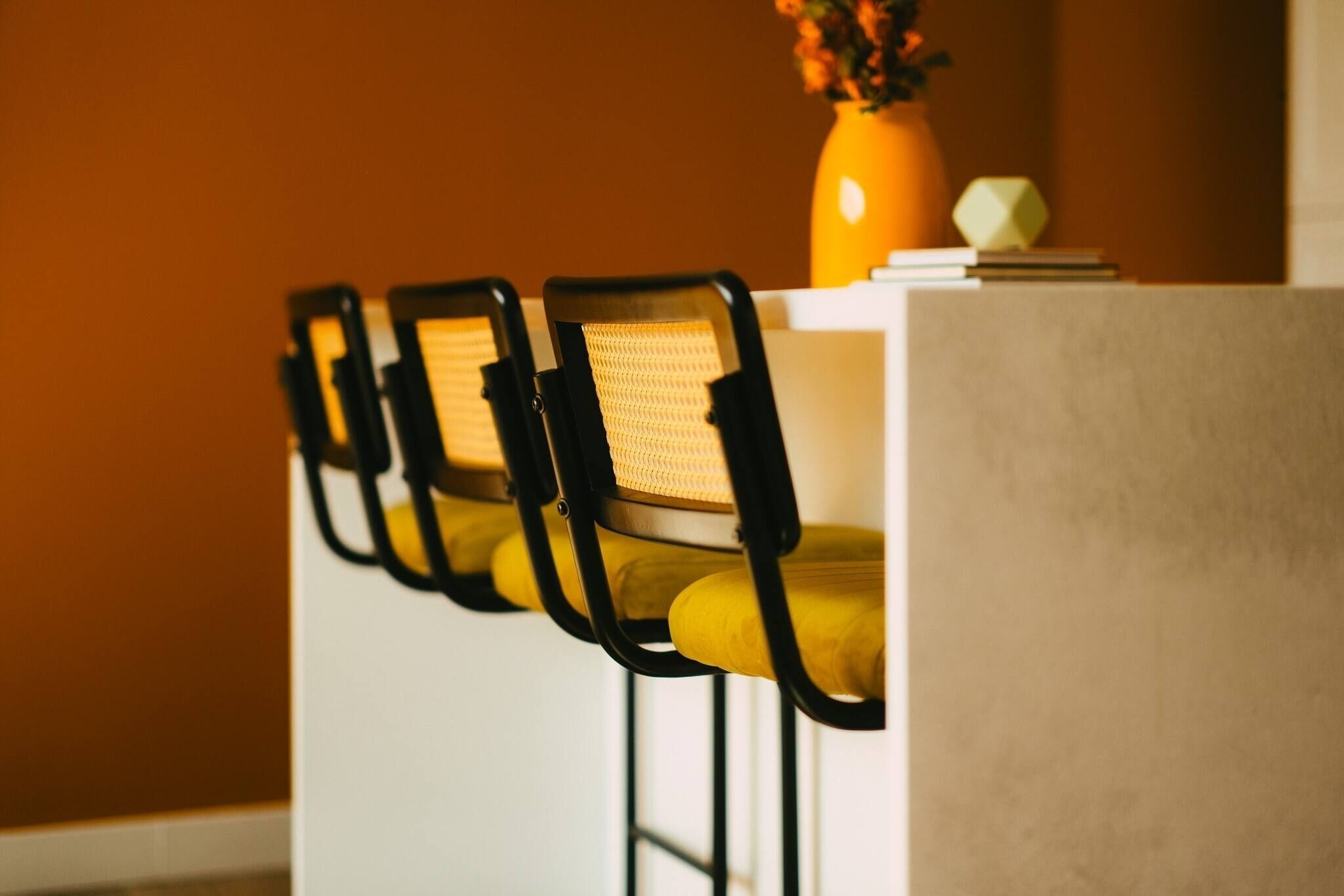A row of chairs with yellow cushions and black frames, a vase with flowers, and a white table against an orange wall.