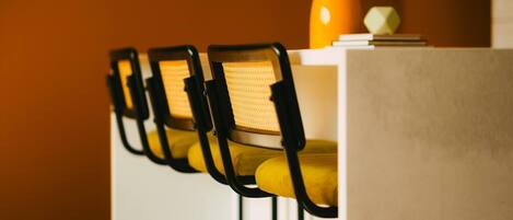 A row of chairs with yellow cushions and black frames, a vase with flowers, and a white table against an orange wall.