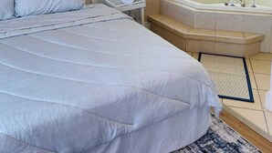 Ένα υπνοδωμάτιο με ένα μεγάλο κρεβάτι, ένα κομοδίνο, ένα φωτιστικό τοποθετημένο στον τοίχο, έναν πίνακα και μια μπανιέρα σε μια γωνία.