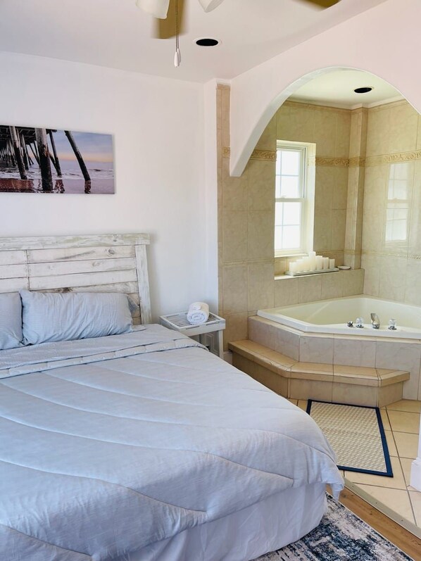 A bedroom with a large bed, a nightstand, a wall-mounted lamp, a painting, and a corner bathtub.