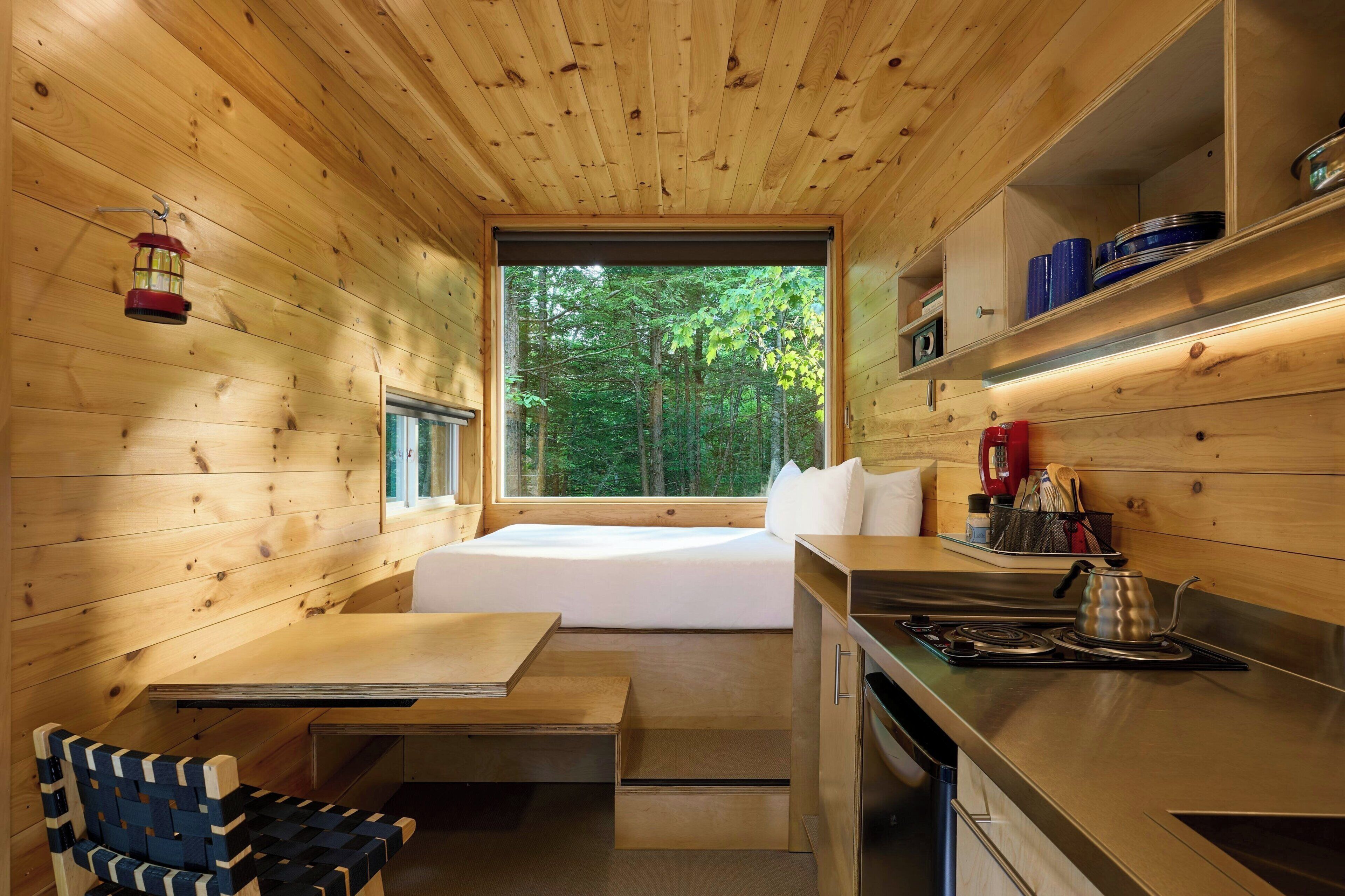 Ein gemütlicher, holzgetäfelter Raum mit Bett, kleinem Tisch, Sitzbank, einer Küchenzeile und einem Fenster mit Blick auf einen Wald.