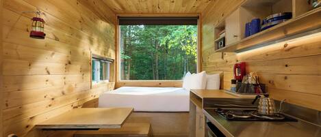 Ein gemütlicher, holzgetäfelter Raum mit Bett, kleinem Tisch, Sitzbank, einer Küchenzeile und einem Fenster mit Blick auf einen Wald.