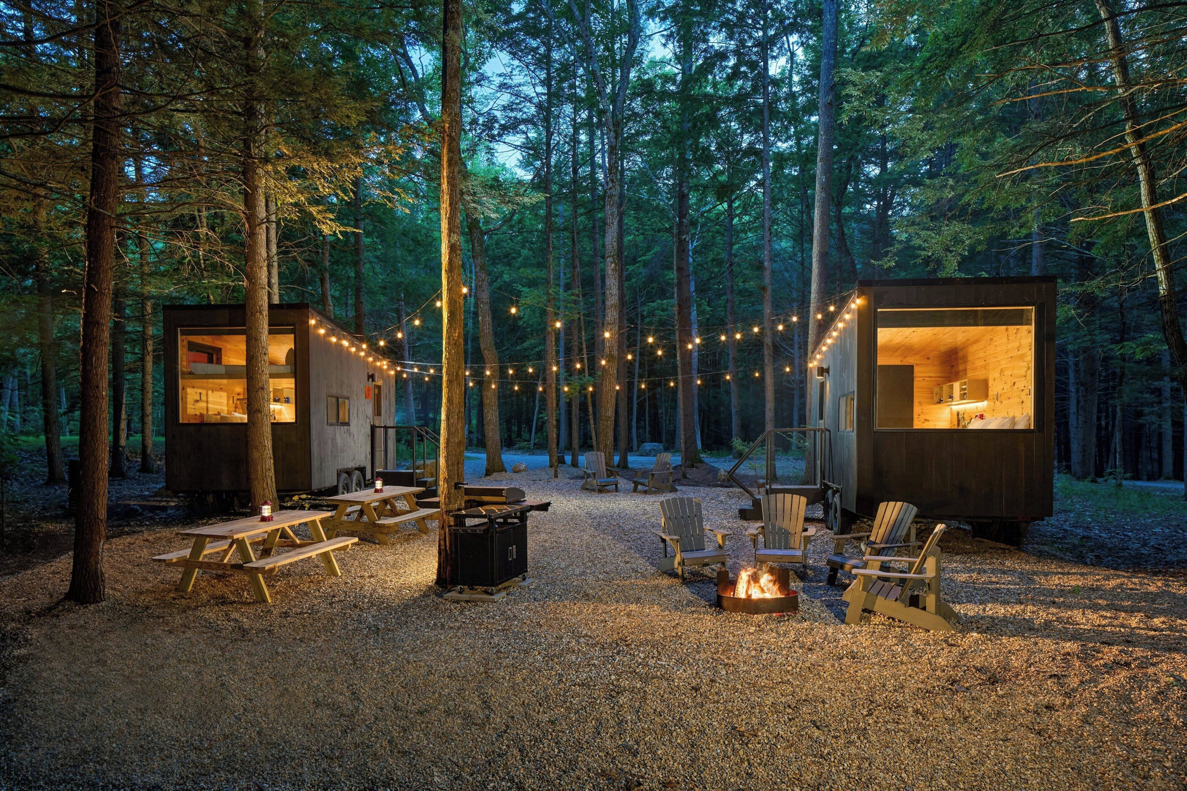 Dos cabañas de madera con luces colgantes, mesas de picnic y un brasero en un bosque.