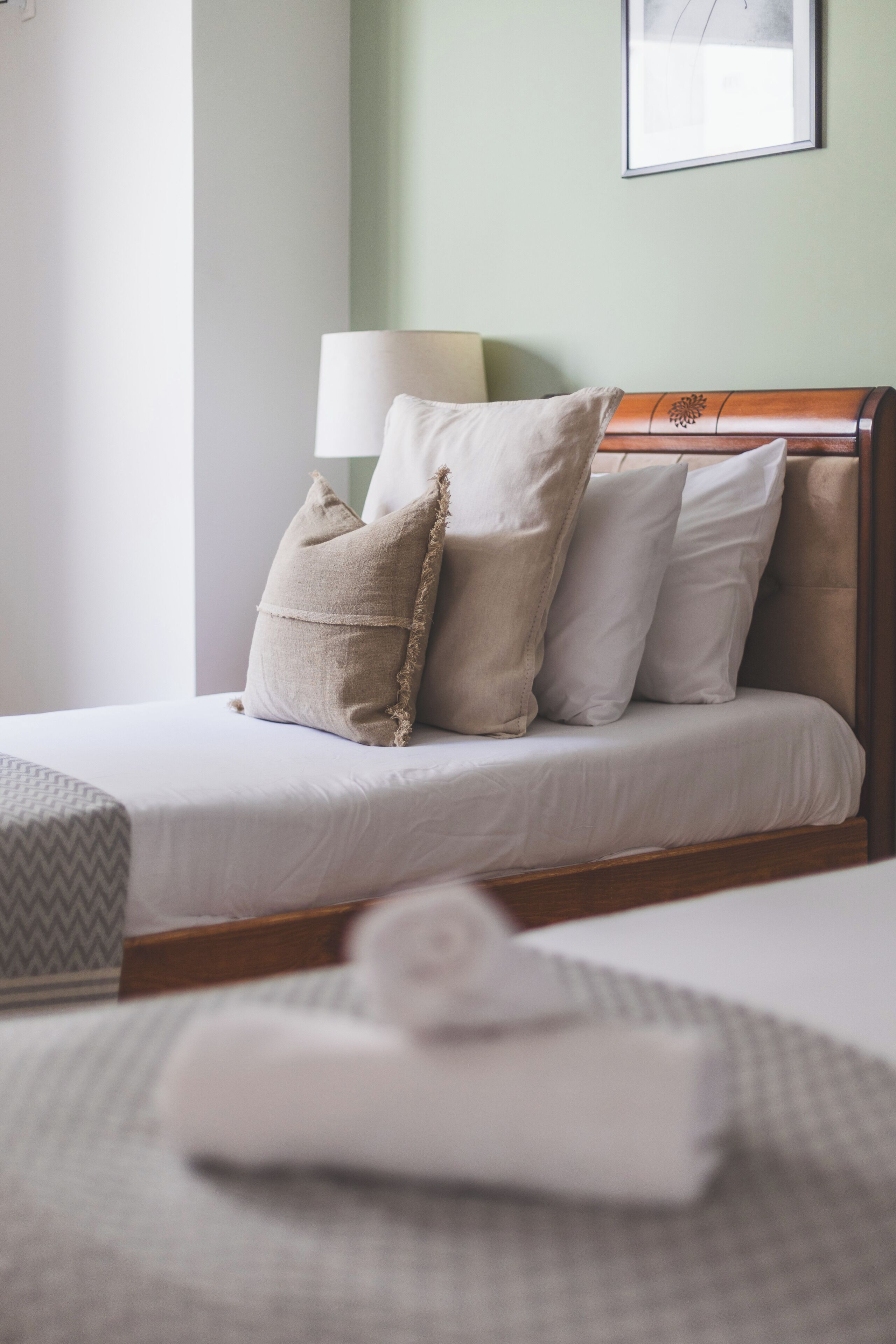 Una cama bien tendida con cabecera de madera, una lámpara y una toalla enrollada sobre una mesita de noche.