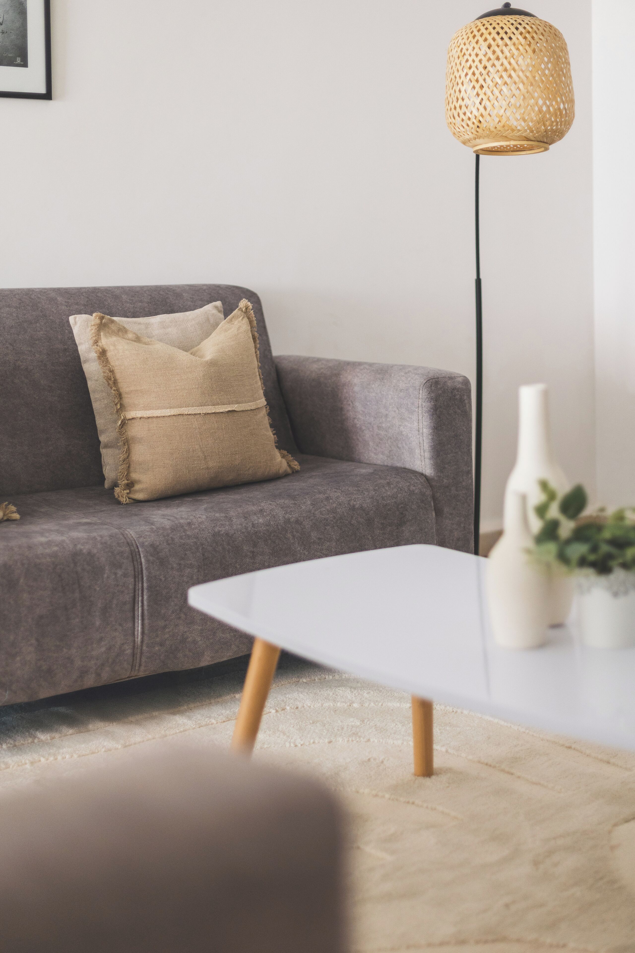 Un canapé gris avec un coussin beige, une table blanche ornée d’un vase et d’une plante, et un grand lampadaire avec un abat-jour en osier.