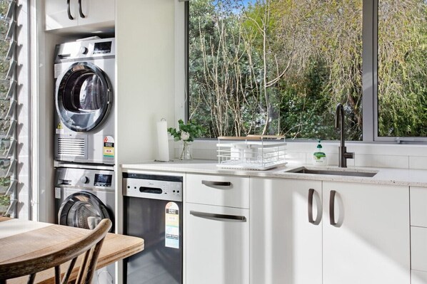 Cocina compacta con lavadora y secadora integradas, lavavajillas y ventana con vistas a árboles.