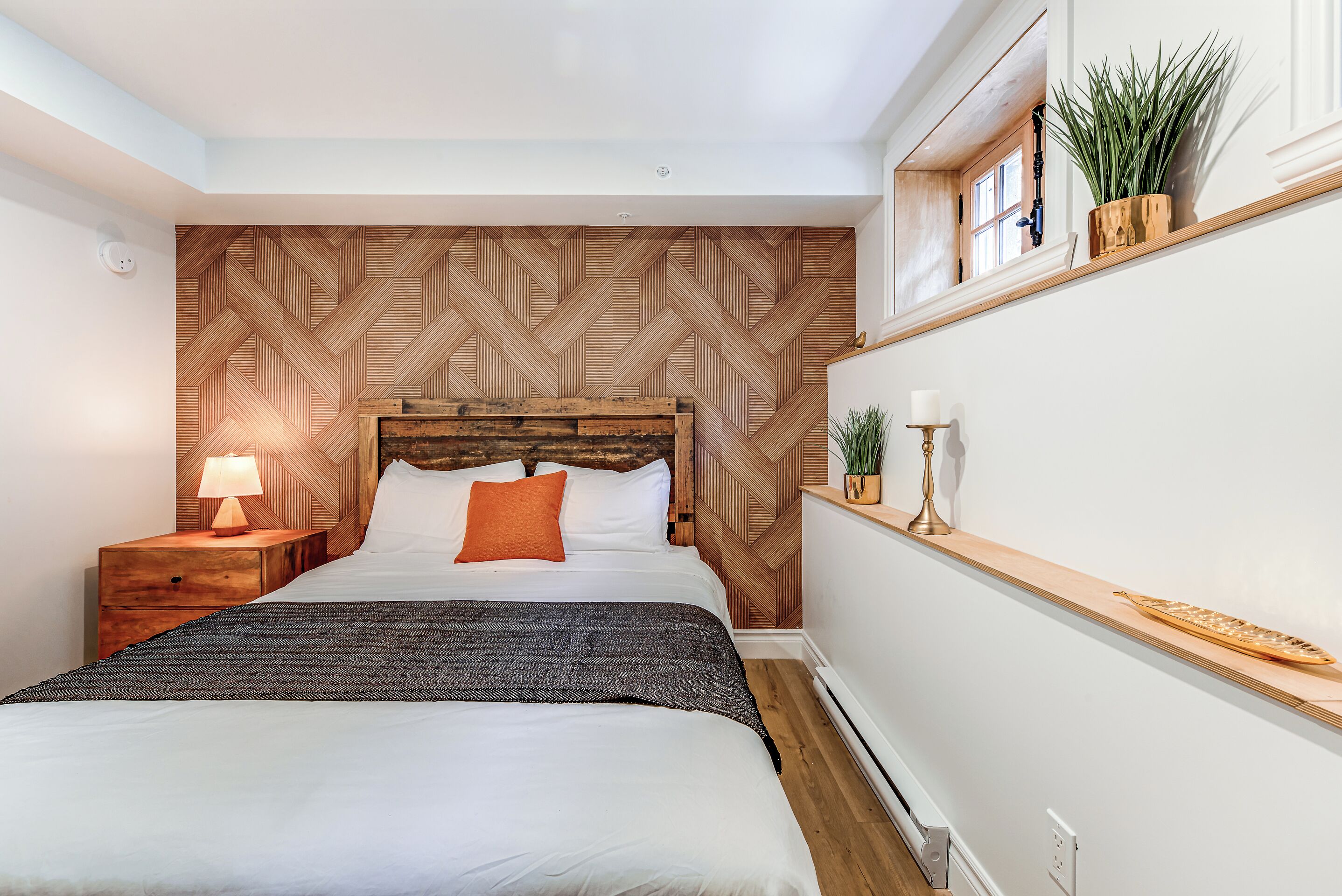 A neatly arranged bedroom with a wooden herringbone-patterned headboard, a bedside table with a lamp, a shelf with decorative items, and a potted plant.