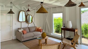 Un salon avec un canapé, une table basse et un fauteuil en osier. On y trouve des lampes suspendues et un miroir au mur.
