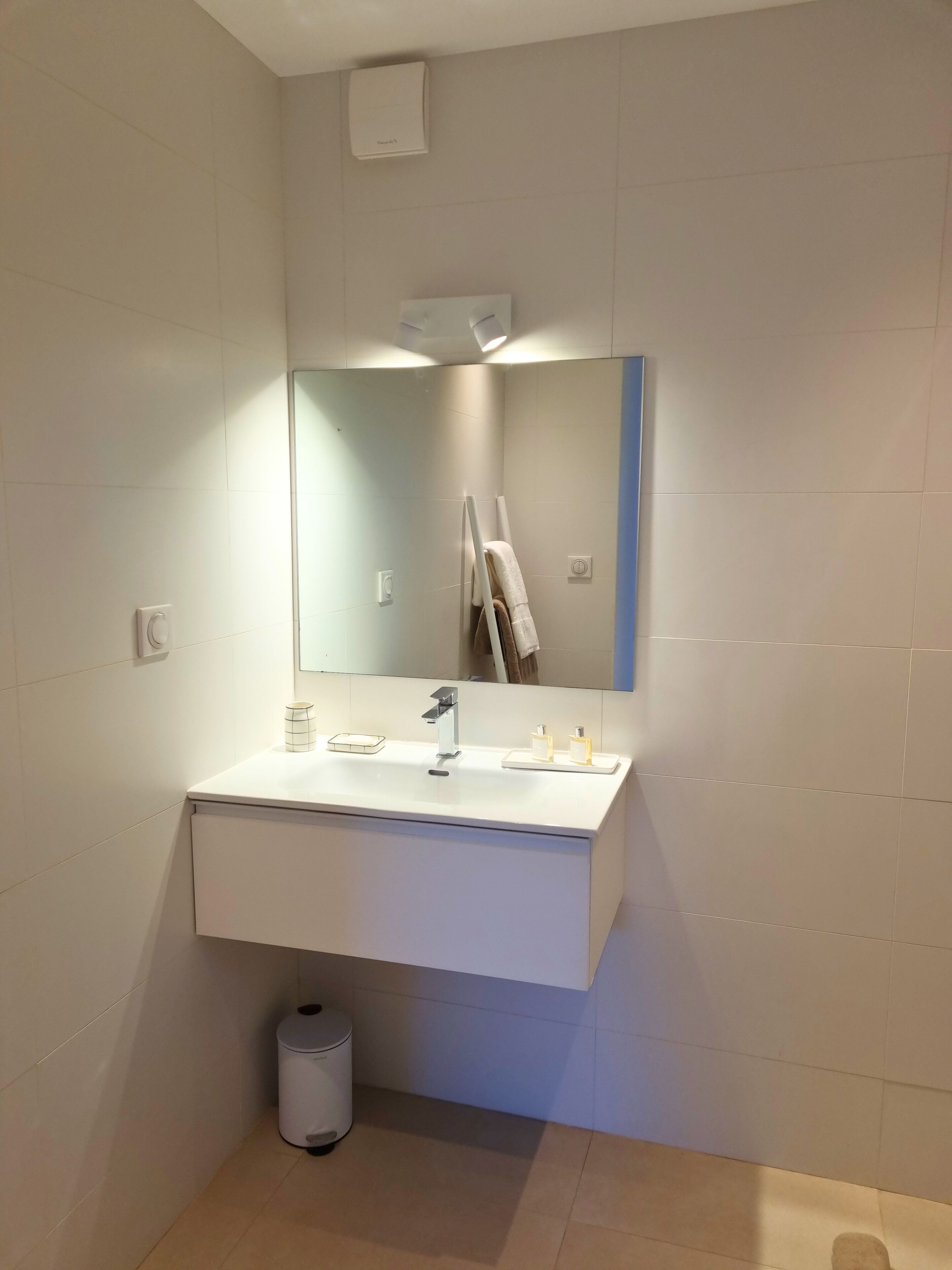 A modern bathroom with a white vanity, a mirror, and a small trash bin.