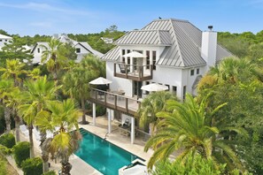 A two-story house with a swimming pool, surrounded by palm trees and other greenery.