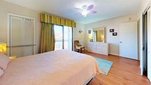Une chambre à coucher comprenant un lit, une commode, une chaise, un ventilateur de plafond et une fenêtre avec des rideaux.