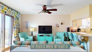 A living room with teal furniture, a ceiling fan, a flat-screen TV, and a sign that reads "THE BEACH IS WHERE MY HEART IS."