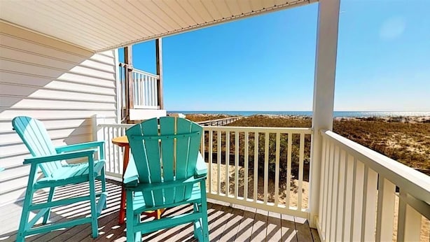A balcony with two turquoise Adirondack chairs and a small table, overlooking a beach with dunes and the ocean.