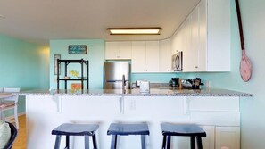 Una cucina con mobili bianchi, un frigorifero in acciaio inossidabile, una macchina da caffè e una pagaia appesa. Di fronte al bancone ci sono tre sgabelli.