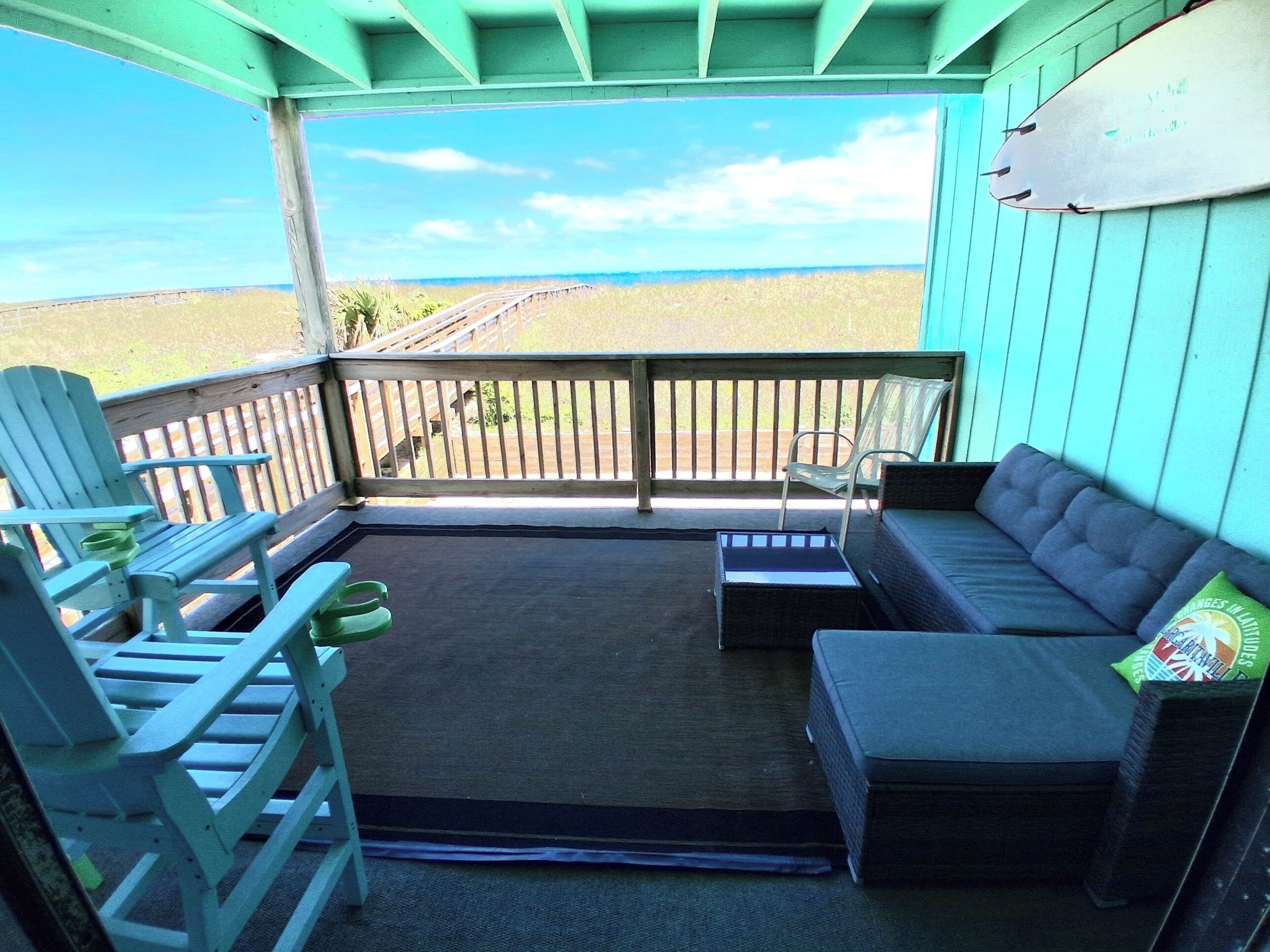 A covered outdoor seating area with a view of the beach, a wooden deck, and a surfboard on the wall.