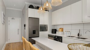 Une cuisine moderne avec des meubles blancs, un réfrigérateur en acier inoxydable, un lave-vaisselle et une machine à café. On y trouve des chaises en osier et un panier tressé sur le comptoir.