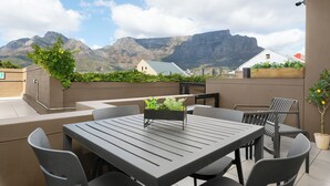 An outdoor patio with a table and chairs, a planter box with plants, and a view of mountains.