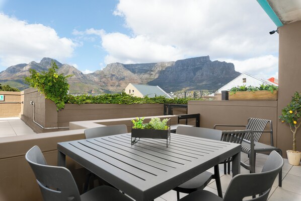 Un patio exterior con mesa y sillas, una jardinera con plantas y vistas a montañas.