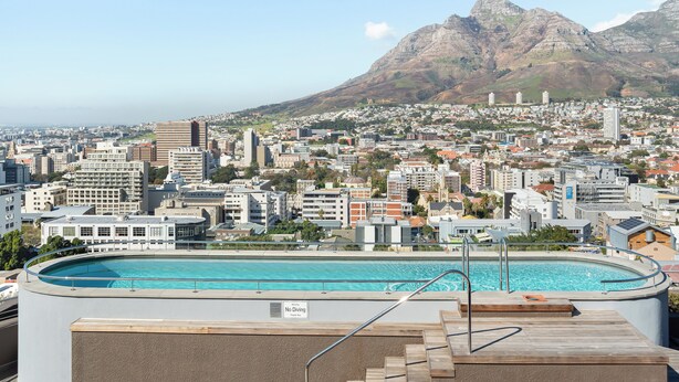 A rooftop pool with a "No Diving" sign, surrounded by a cityscape with buildings and mountains in the background.
