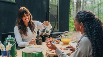 Deux personnes sont assises à une table en bois, devant une machine à café, un bol et une carafe de jus d’orange.