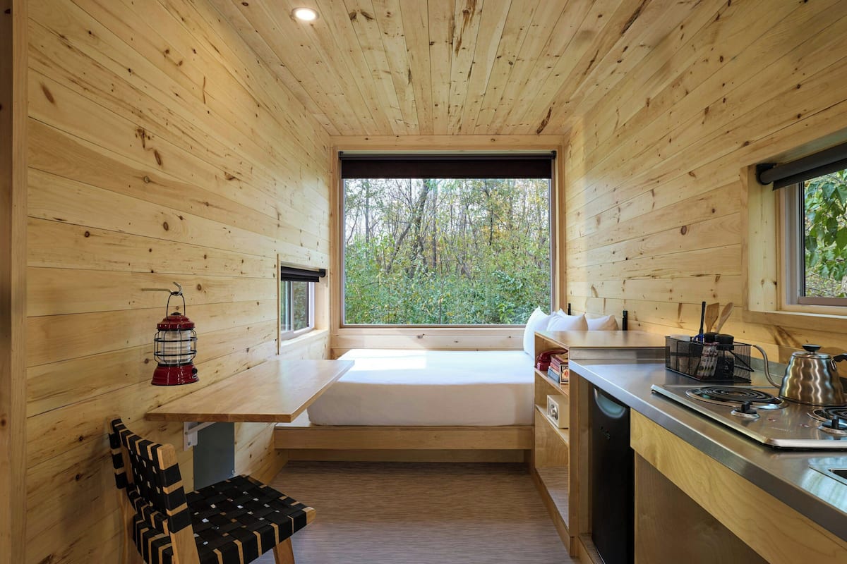 A compact cabin with a wooden interior, a built-in bench, a small table, a window with a view of trees, and a kitchenette area.