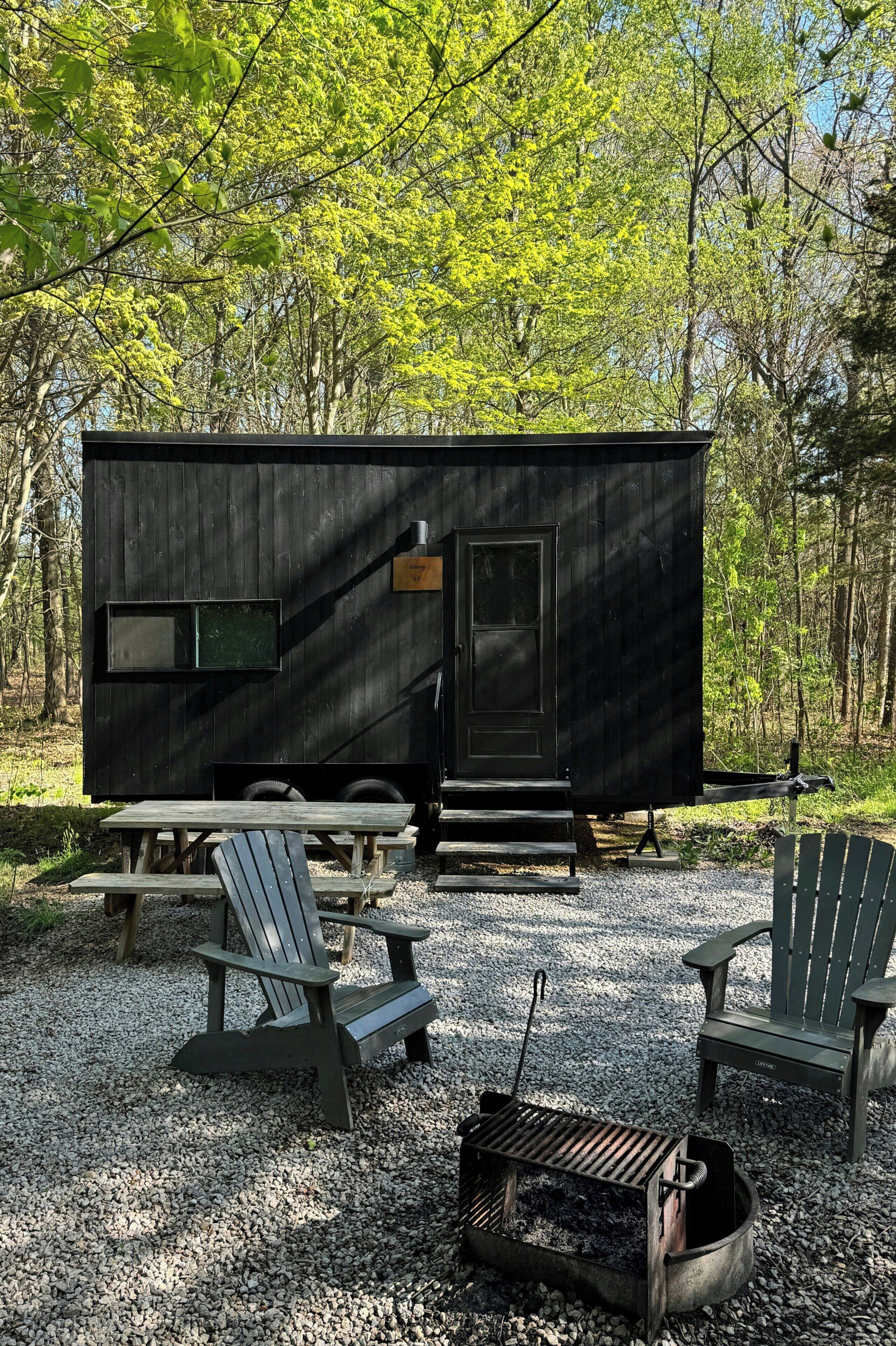 Una caravana negra con una pequeña ventana y puerta, rodeada de grava, mesas de picnic de madera y sillas Adirondack.