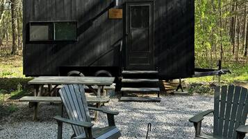 Una caravana negra con una pequeña ventana y puerta, rodeada de grava, mesas de picnic de madera y sillas Adirondack.