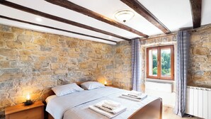 A bedroom with a stone wall, wooden beams, a bed with linens, a nightstand, a window with a view, and a radiator. - Boutique House Rustikana with pool Korte (Izola)