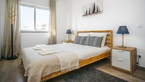 A neatly arranged bedroom with a wooden bed, two bedside tables with lamps, a window with curtains, and a sailboat painting on the wall. - Linaria Apartment, Portimão, Algarve (Portimão)