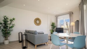 A modern living room with a grey sofa, a round dining table with blue chairs, a large potted bamboo plant, and a decorative wall piece. - Linaria Apartment, Portimão, Algarve (Portimão)