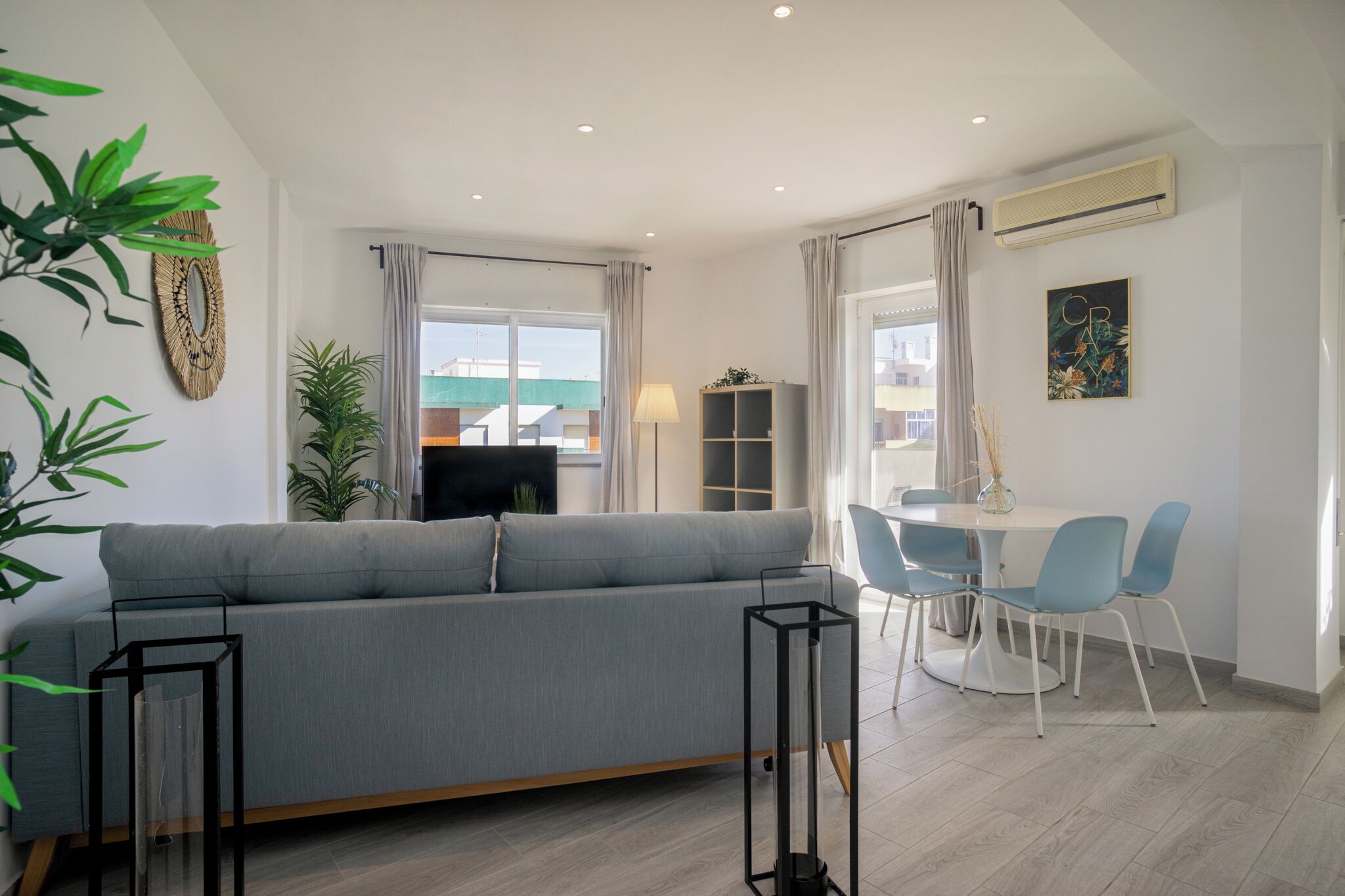 A modern living room with a grey sofa, a dining table with blue chairs, and a bookshelf.
