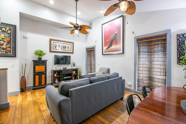 A modern living room with a grey sofa, wooden flooring, a dining table with chairs, a ceiling fan, and framed artwork on the walls.