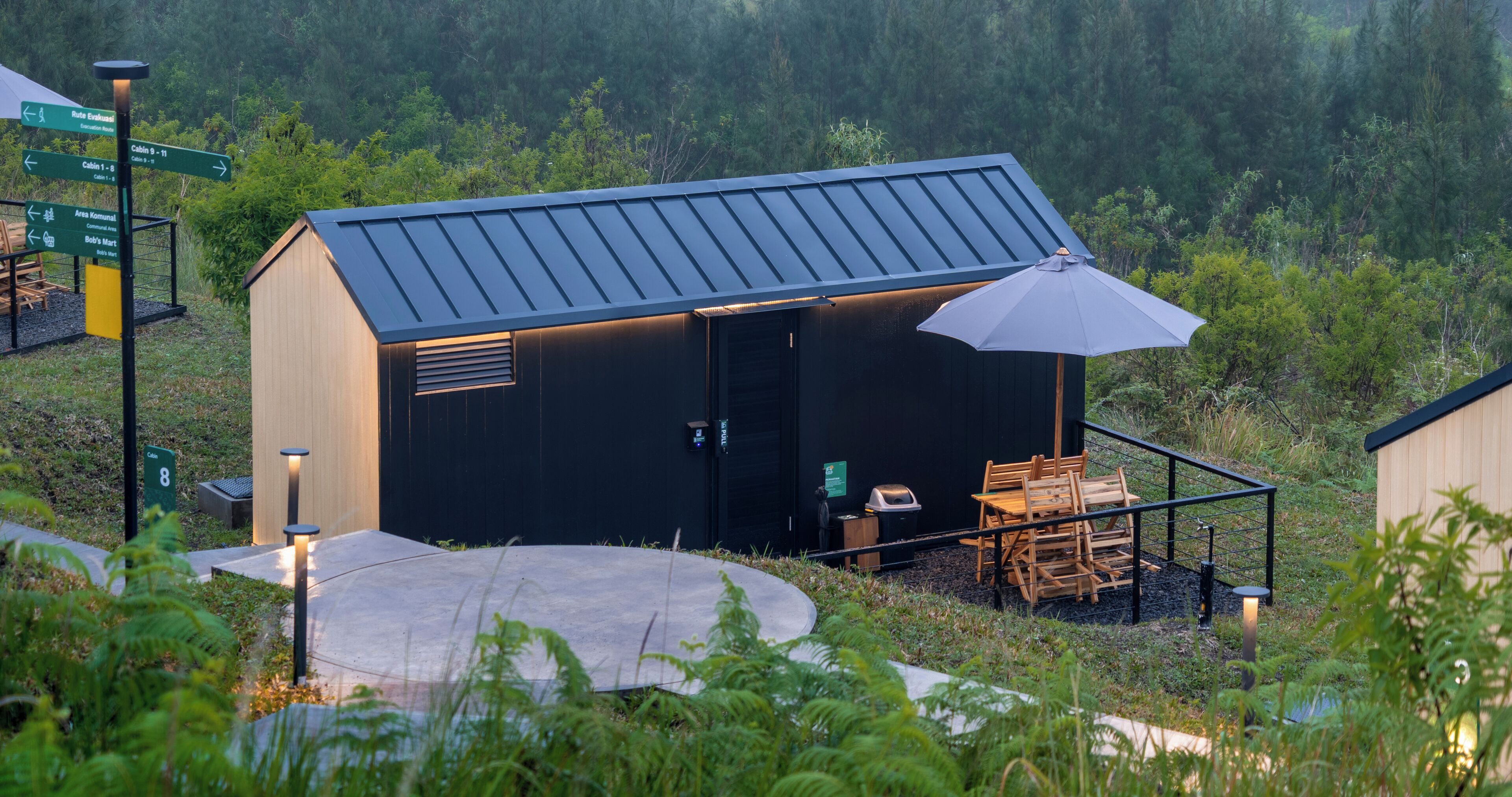 A small, dark-colored building with a metal roof, a patio with chairs, and a table. There are directional signs and a trash bin nearby.