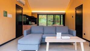 Een moderne hotelkamer met een grijze hoekbank, een witte salontafel en uitzicht op het buitengebied door grote ramen.