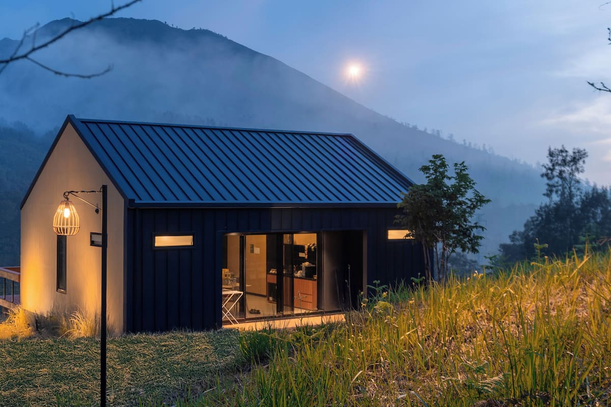 Une maison moderne avec un toit foncé, de grandes fenêtres et un intérieur visible, comprenant un espace repas.