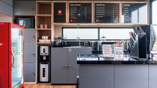 Eine moderne Küchenzeile mit Kaffeemaschine, Wasserspender und Mikrowelle. Regale mit Kartons und eine Speisekarte befinden sich über der Arbeitsplatte.