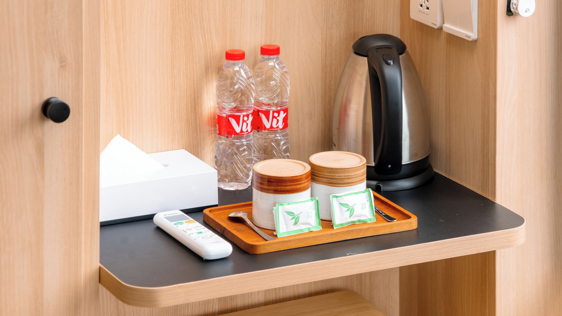 Un mini bar con una botella de agua, una tetera, un control remoto y algunos frascos sobre una bandeja de madera.