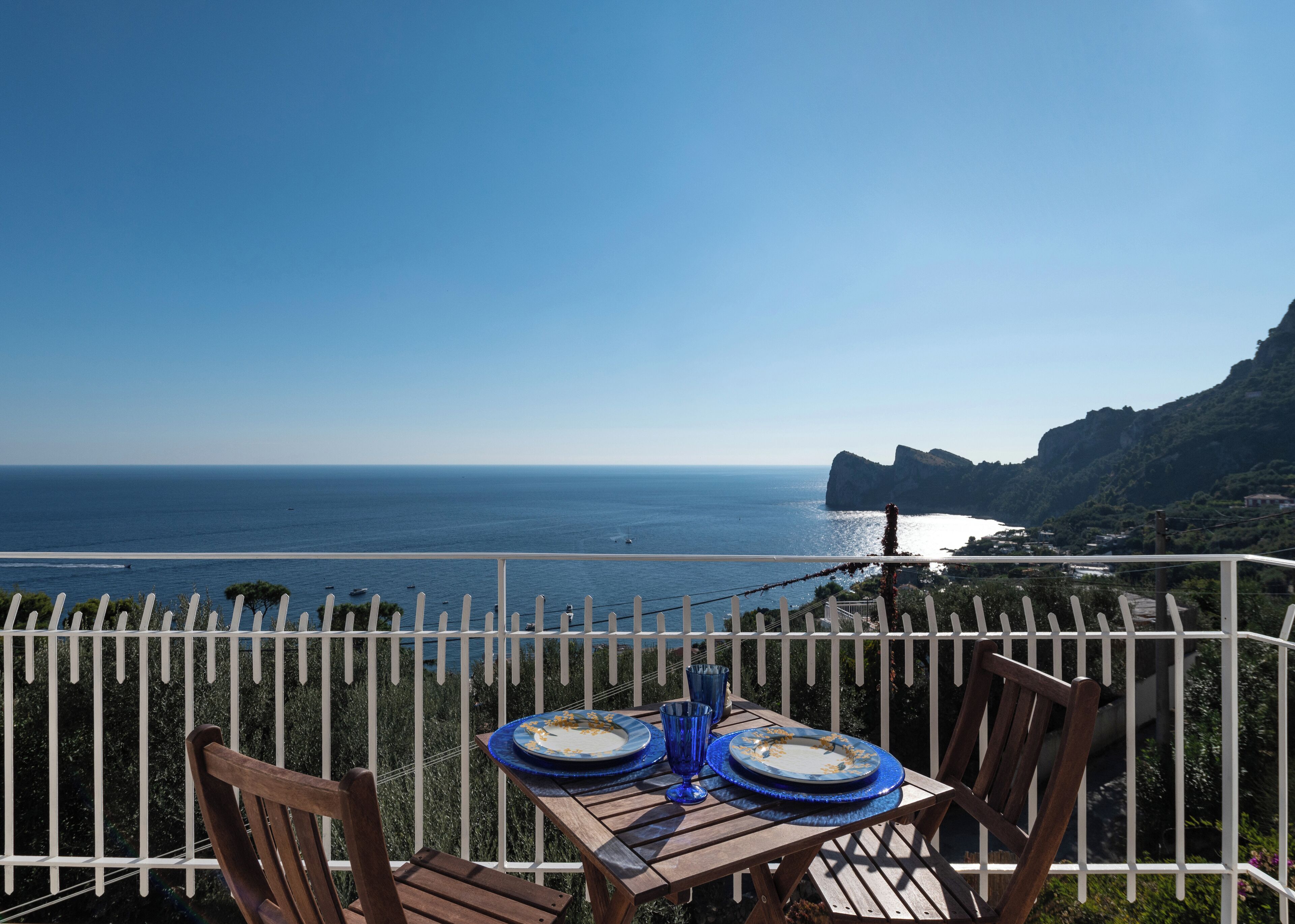 A table with plates and blue glasses on a balcony overlooking a coastal view with a rocky outcrop.