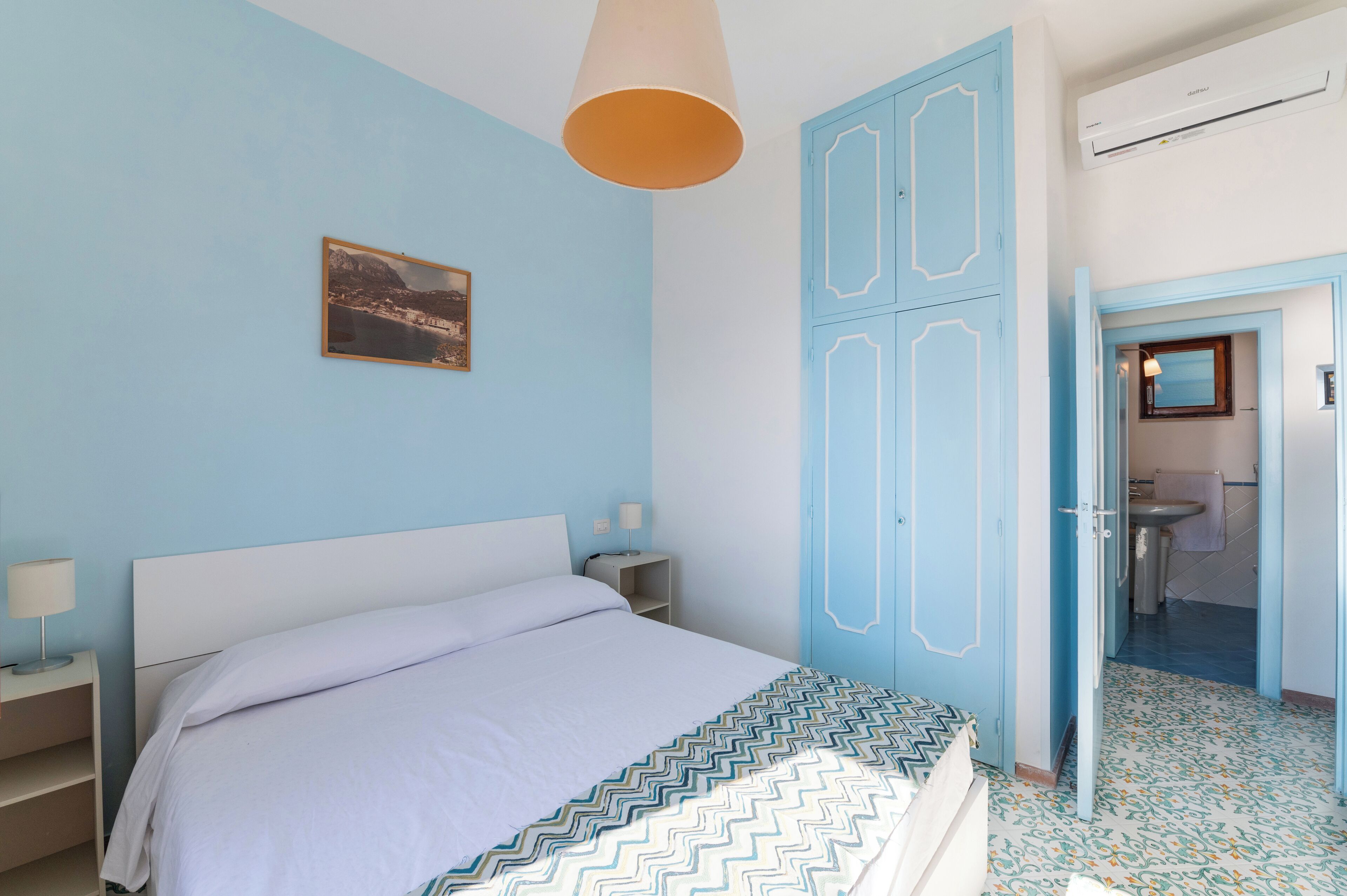 A bedroom with a bed, nightstands, a hanging light, a wardrobe, and a bathroom visible through an open door.