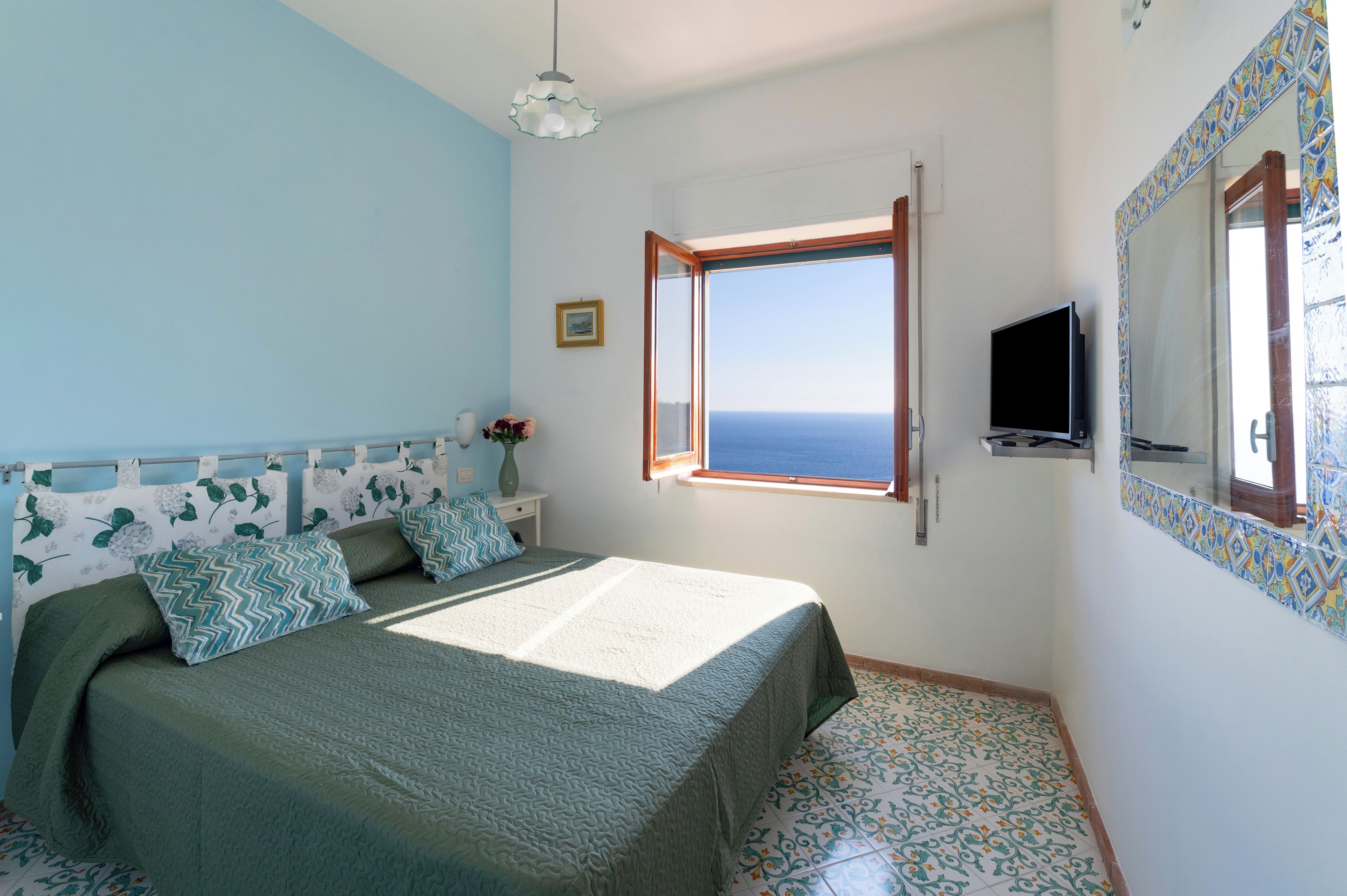 Una camera da letto con un letto, una TV, una finestra vista mare e un pavimento piastrellato.
